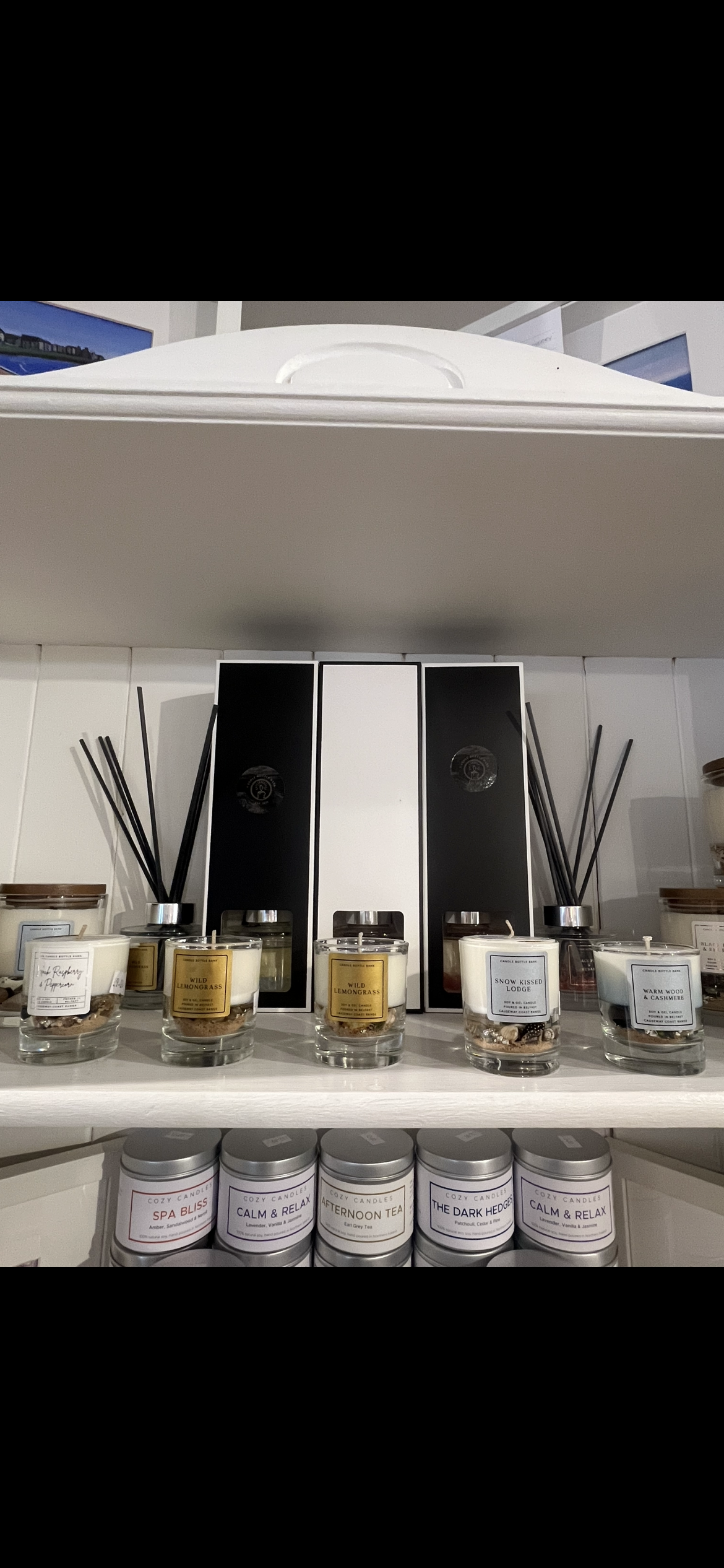 A shelf display of home fragrance products, including reed diffusers with black reeds in clear glass jars and two tall rectangular boxes, alongside several small glass jars with silver lids. The products are arranged on white shelves against a white tiled wall.
